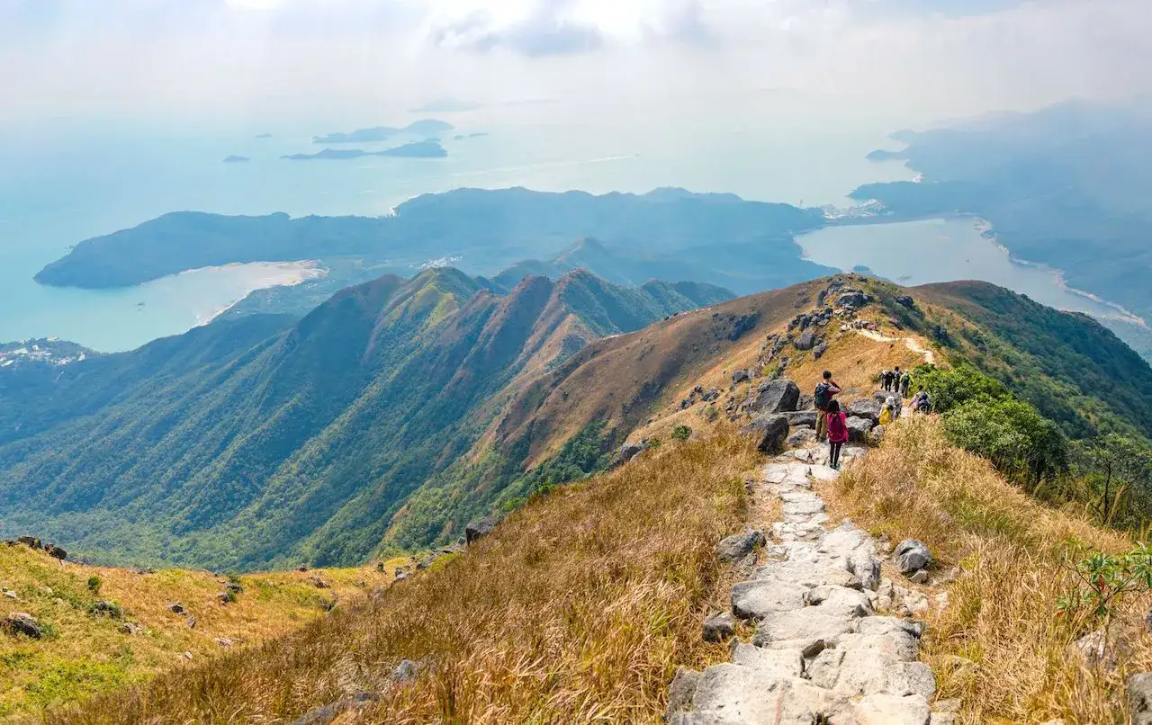 Wanderer genie&szlig;en eine atemberaubende Aussicht bei einer Wanderung in Hongkong. Der steinige Pfad schl&auml;ngelt sich &uuml;ber gr&uuml;ne Berge zum glitzernden Meer.