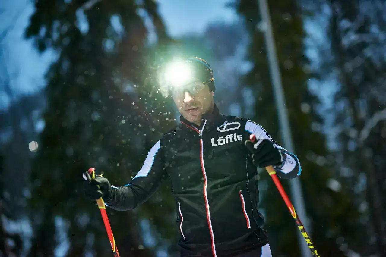 Ein Mann mit Stirnlampe l&auml;uft im Winter mit Skist&ouml;cken durch verschneiten Wald.
