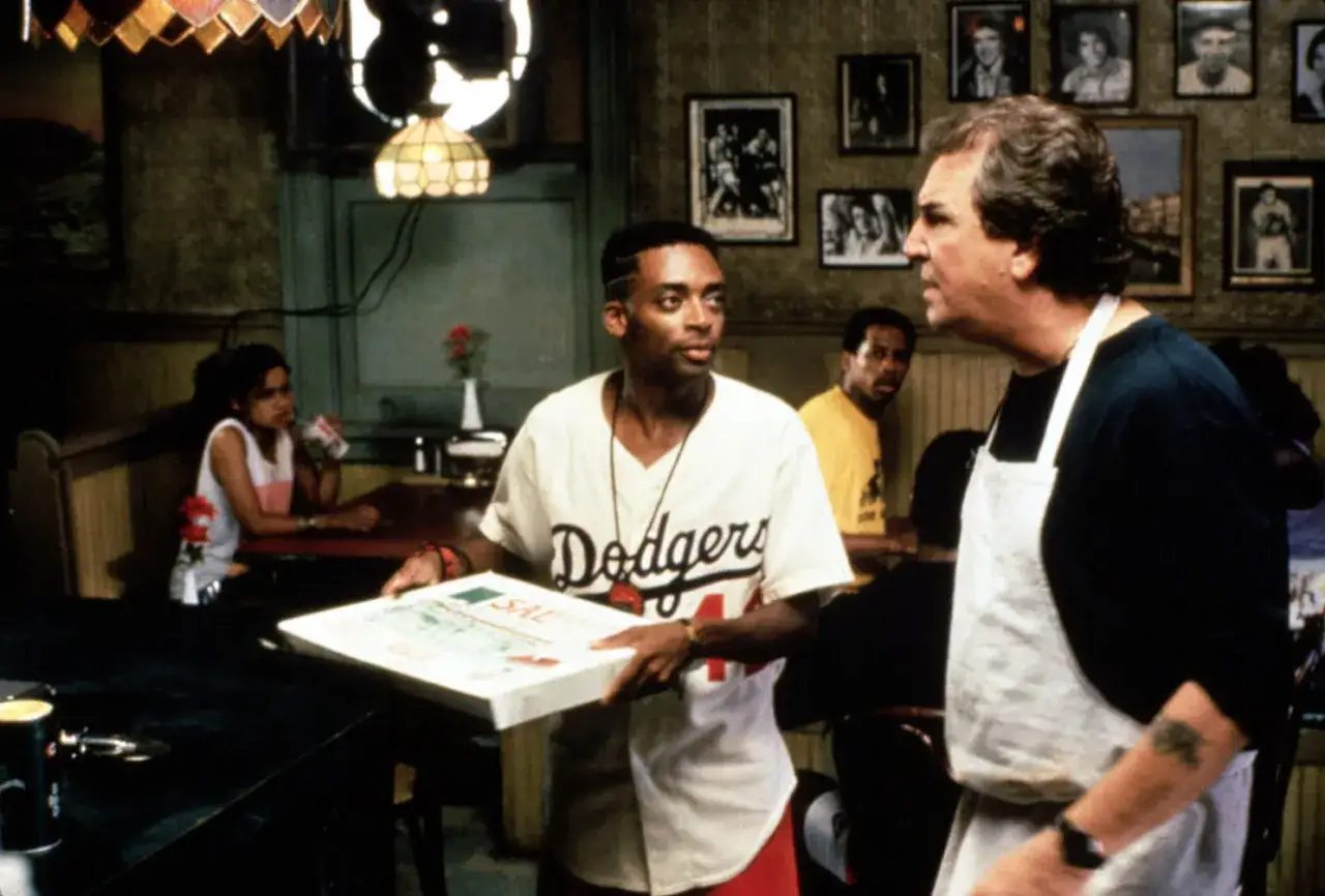 Spike Lee w filmach, trzymający pudełko z pizzą, rozmawia z mężczyzną w fartuchu.
