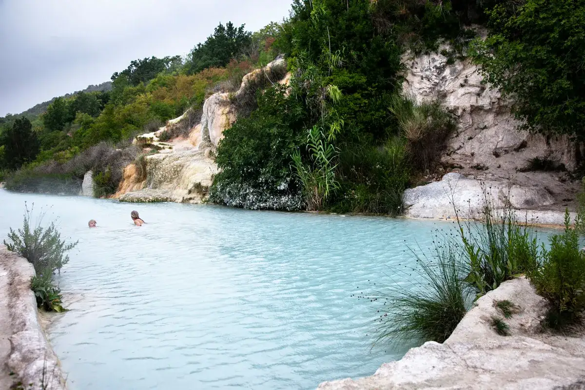 Bagno Vignoni: cascate naturali con acqua azzurra e vegetazione lussureggiante. Due persone nuotano nelle terme.