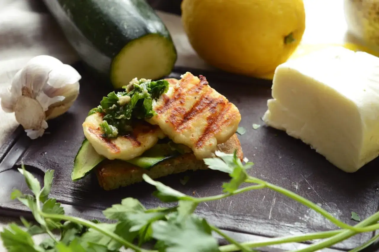 Grillowany ser halloumi na grzance z cukinią i ziołami. Obok czosnek, cytryna i kawałek sera.