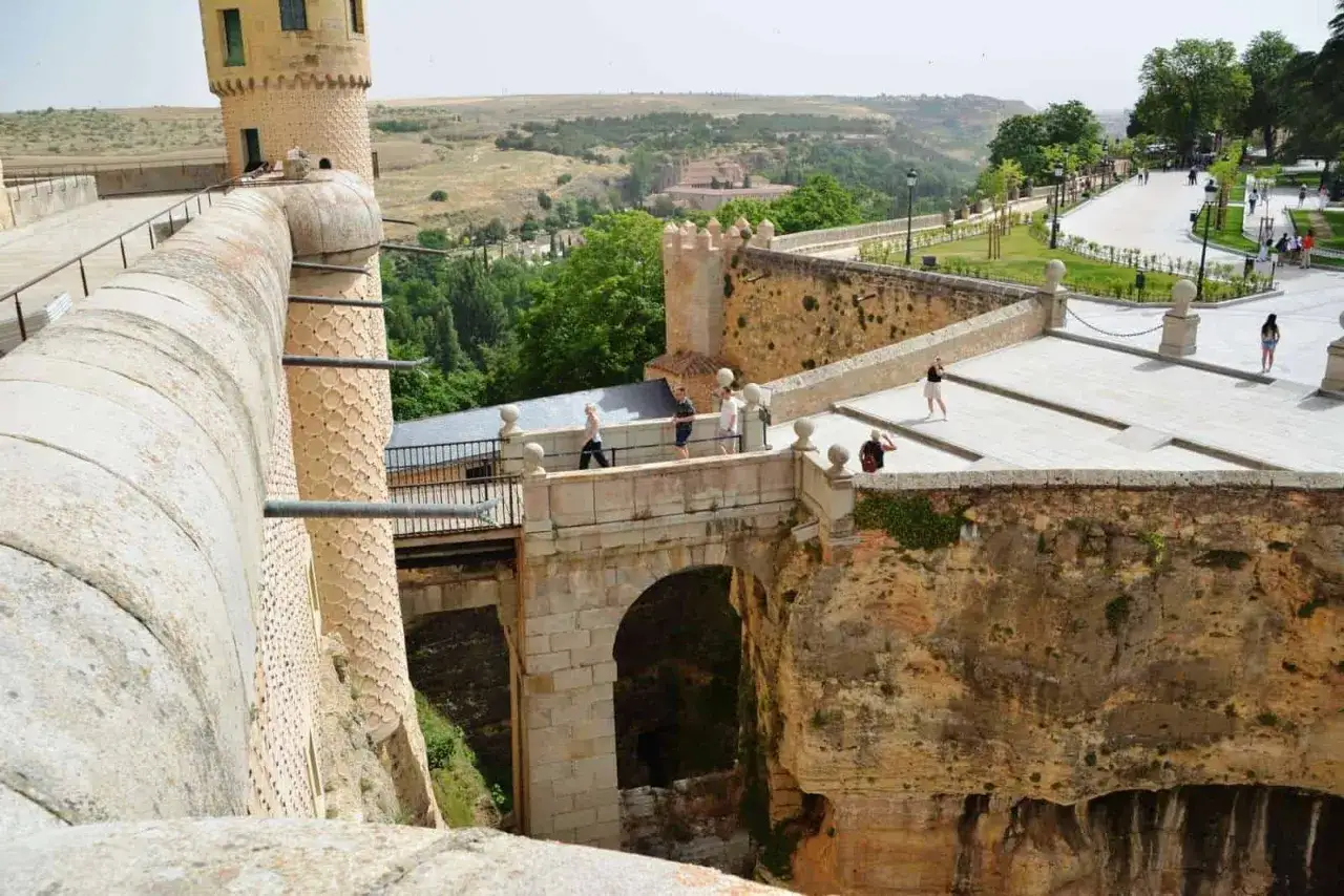 El **castillo de Segovia**, con sus imponentes murallas y torres, se alza sobre un paisaje verde. Turistas pasean por sus puentes y terrazas, disfrutando de las vistas.