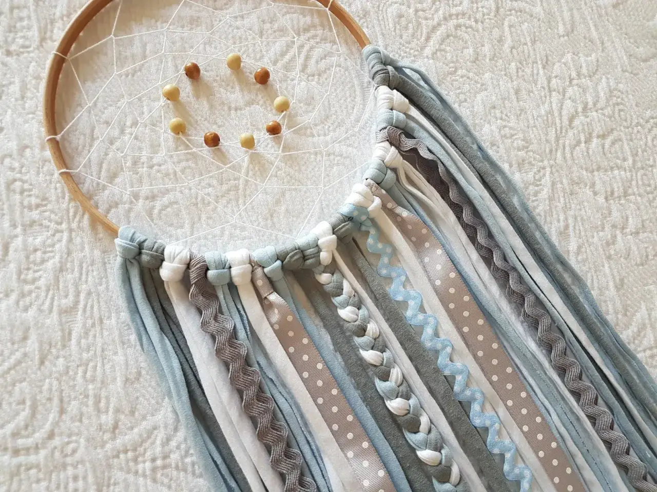 Un atrapasue&ntilde;os casero con aros de madera, red blanca y cuentas. Cintas de tela y encaje cuelgan en tonos azules, grises y blancos.