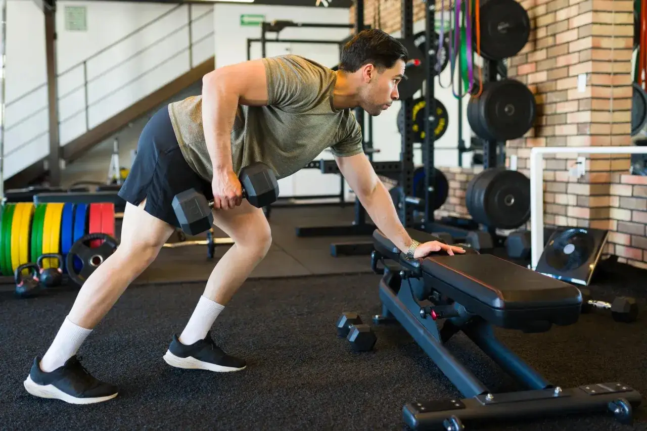 Homme faisant un rowing haltère, soutenu par un banc de musculation dans une salle de sport.
