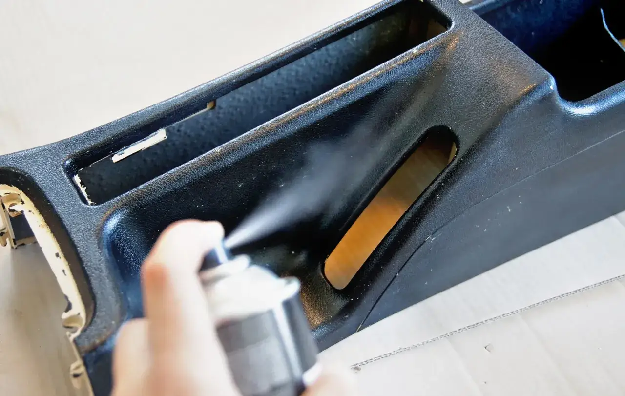 Ręka rozpyla środek na czarny plastikowy element konsoli środkowej auta, pokazując jak odnowić plastiki w aucie.