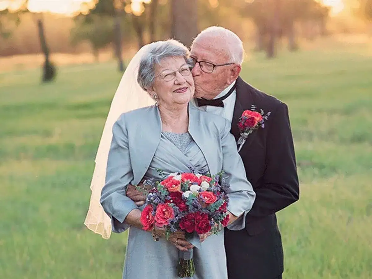Pareja mayor celebra sus bodas de oro. Él le da un beso en la mejilla mientras ella sonríe, sosteniendo un ramo. ¡Un discurso gracioso para bodas de oro sería perfecto para ellos!