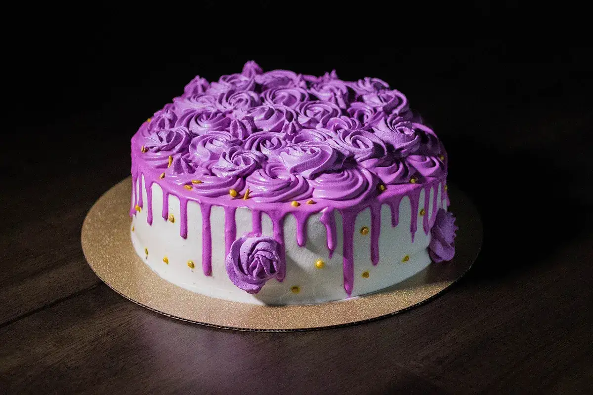 Torta decorata con abbondante crema al burro viola a forma di rose, glassa rosa che cola e perline dorate.