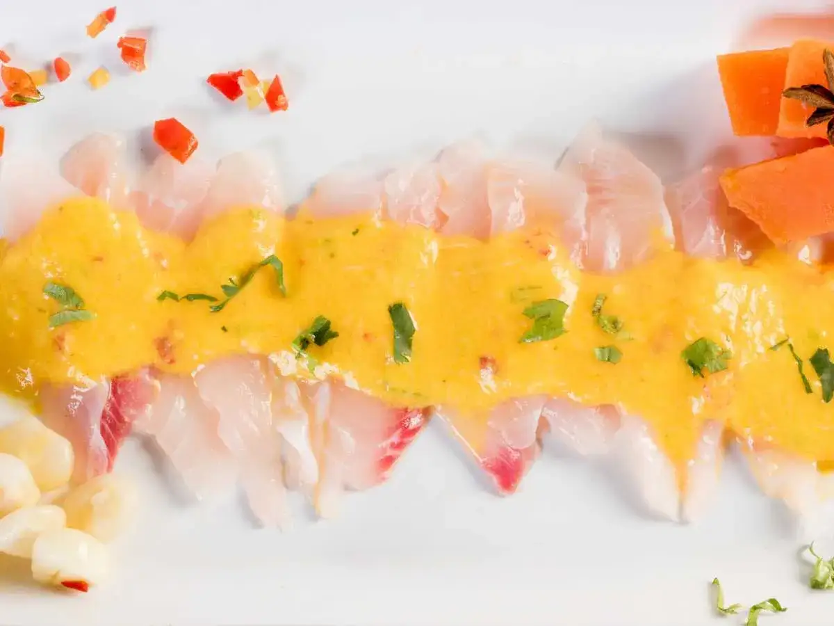 Delicioso tiradito de corvina con salsa de ají amarillo, adornado con choclo, pimientos y camote.
