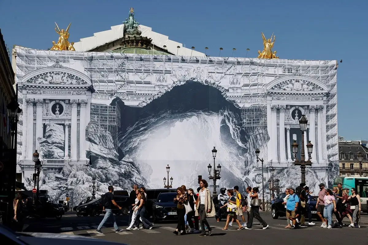 Des passants traversent une rue devant l'Op&eacute;ra Garnier, dont la fa&ccedil;ade est recouverte d'une b&acirc;che monumentale illustrant des travaux artistiques.