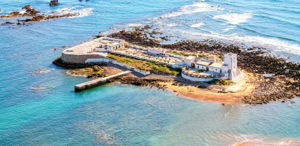 Festung Sancti Petri auf einer felsigen Insel im t&uuml;rkisfarbenen Meer, mit einem Steg und einem kleinen roten Boot.