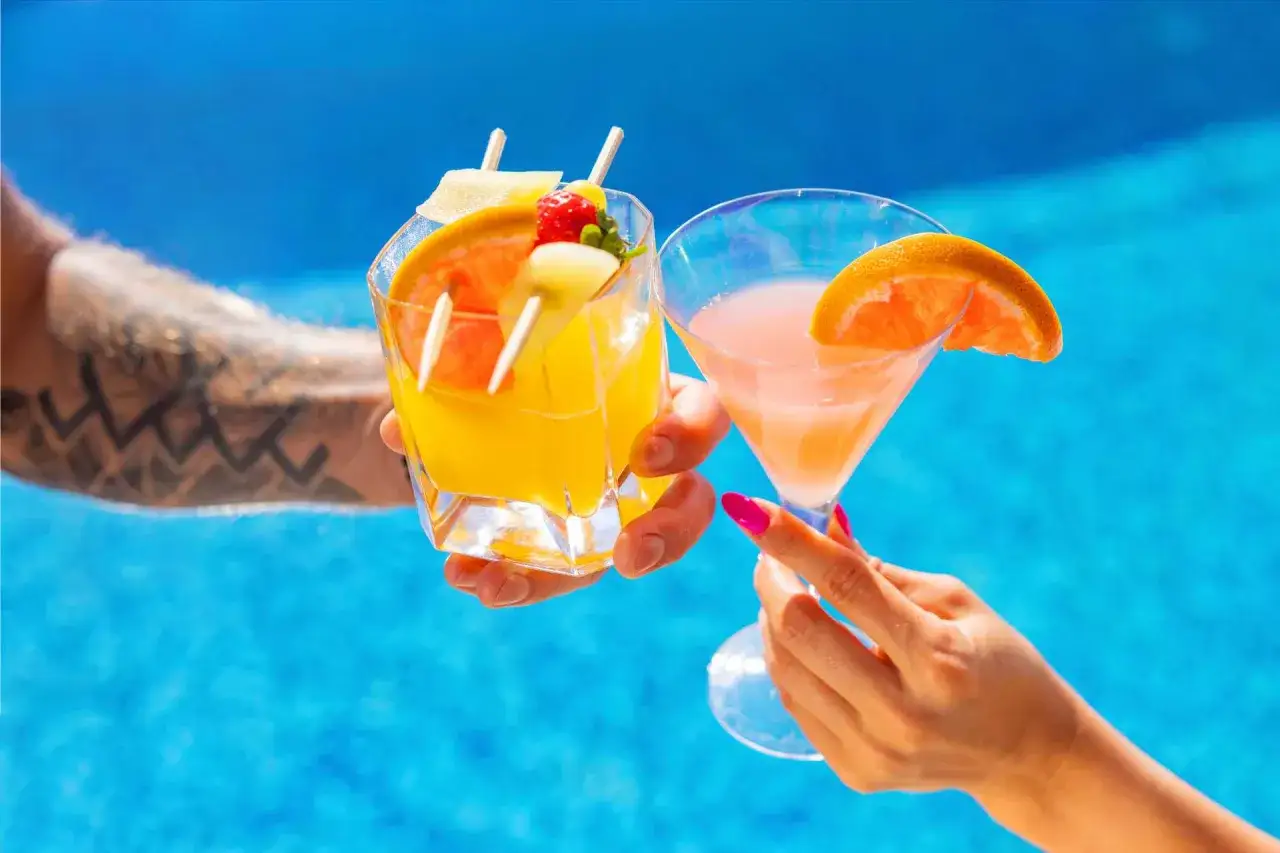 Dwie ręce unoszą kolorowe drinki z owocami nad basenem. Idealny moment na wakacje all inclusive soft.