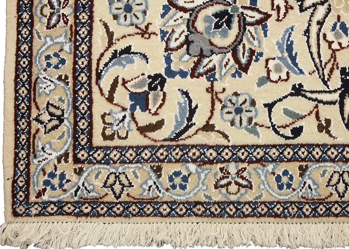 Dettaglio di un tappeto persiano più pregiato con motivi floreali intricati su sfondo crema, bordi blu e frange.