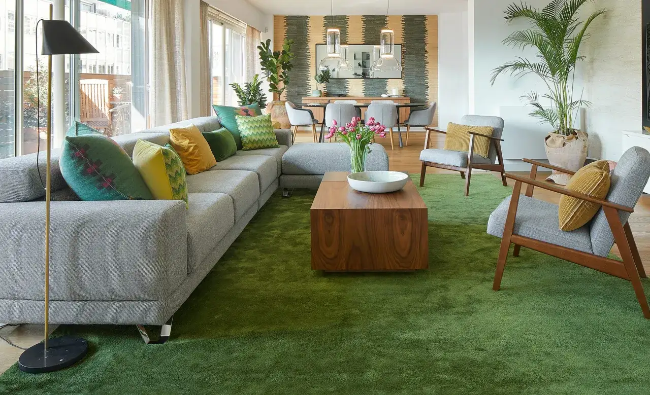 Sof&aacute; modular gris con cojines verdes y amarillos, alfombra verde vibrante y mesa de centro de madera. Muebles pintados en verde que crean un ambiente fresco.