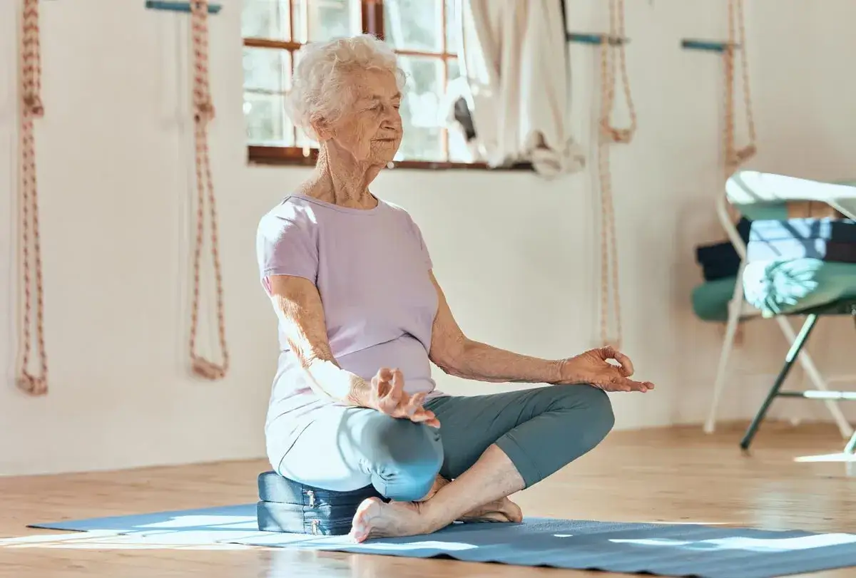 Une femme &acirc;g&eacute;e m&eacute;dite, les yeux ferm&eacute;s, dans une salle de yoga. Une pratique b&eacute;n&eacute;fique pour la stimulation cognitive.