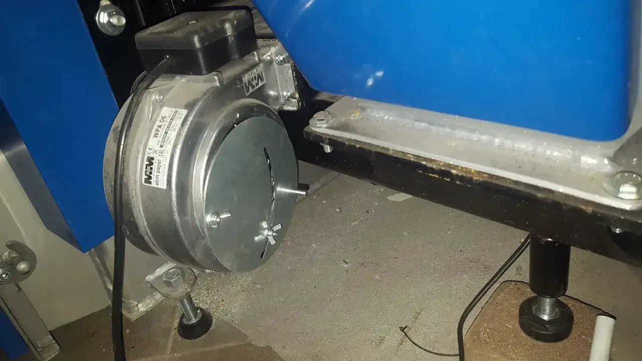 Metalowa dmuchawa do pieca na ekogroszek, model WPA 06, zamontowana obok niebieskiego zbiornika.