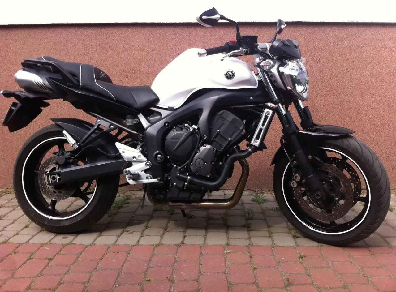 Biały motocykl Yamaha FZ6 z czarnym siedzeniem i detalami, zaparkowany na czerwonej kostce brukowej przy brązowej ścianie.