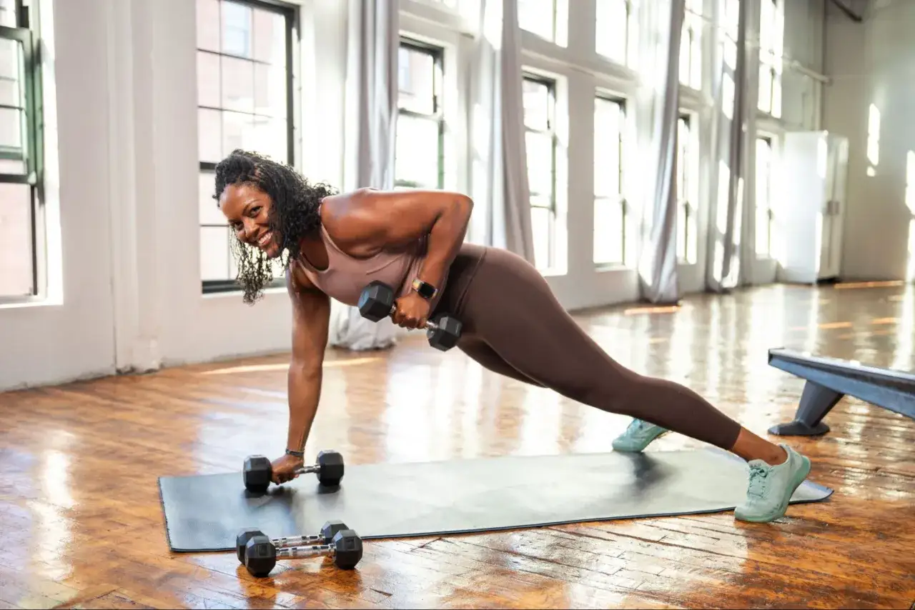 Donna sorridente esegue un rematore con manubri in posizione di plank. Scopri cos'&egrave; il circuit training per un allenamento completo.