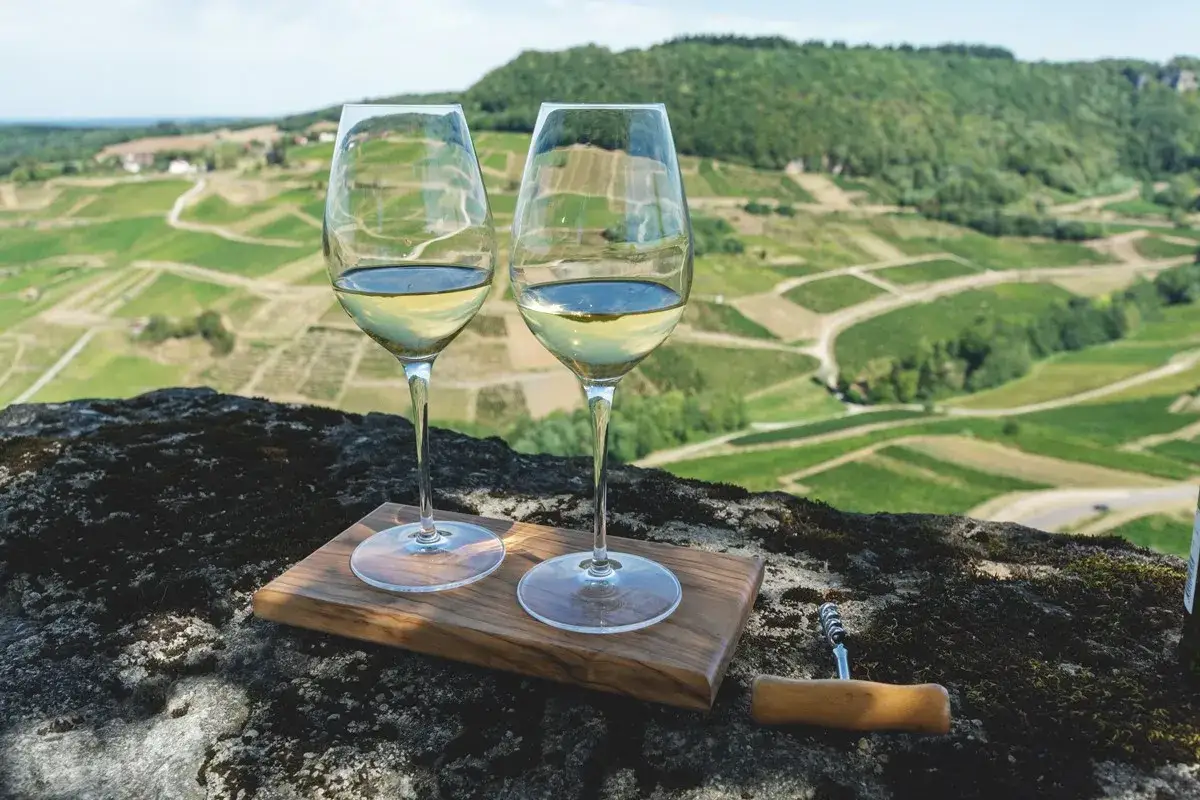 Due calici di vin jaune dorato, su un tagliere in legno, con un paesaggio di vigneti a terrazze sullo sfondo.