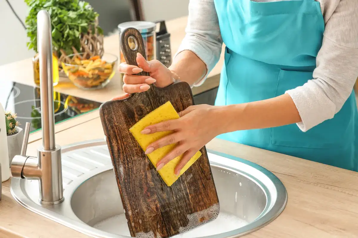 Frau reinigt ein Holz-Schneidebrett mit einem gelben Schwamm in der Spüle. Backpulver hilft, hartnäckige Flecken zu entfernen.