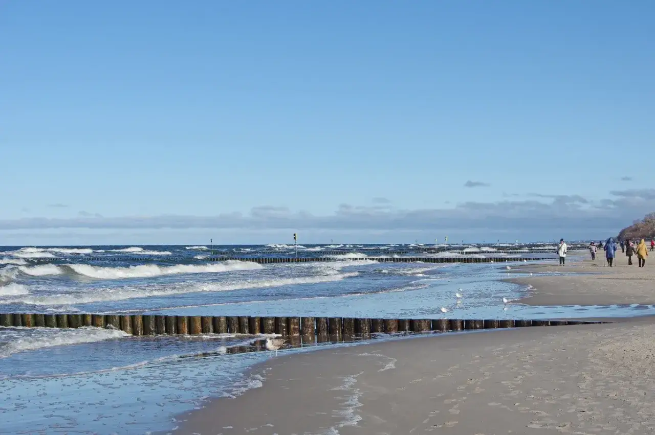 Spacer po plaży, fale rozbijające się o drewniane falochrony. W tle ludzie spacerujący. Idealne miejsce na relaks, jak w