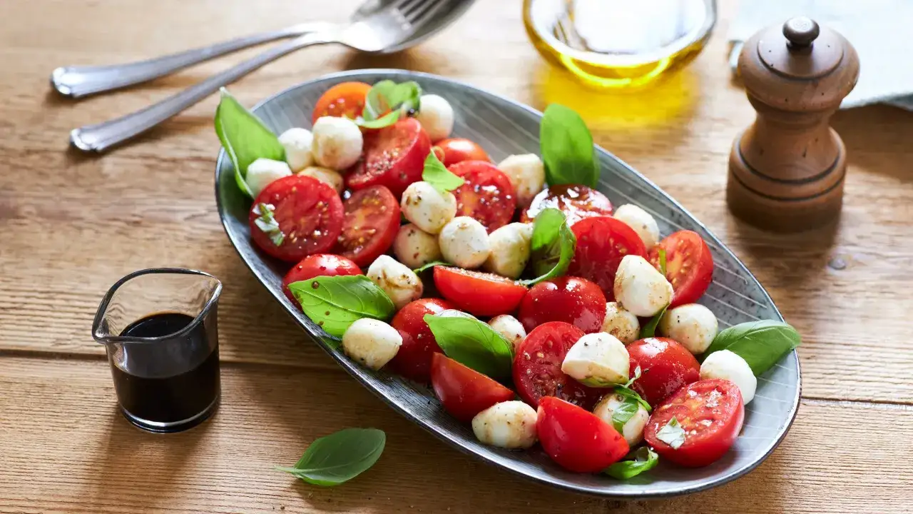 Frischer Tomate Mozzarella Salat mit Basilikum, serviert auf einem ovalen Teller. Daneben ein kleines Glas Balsamico und ein Pfeffermühlen.