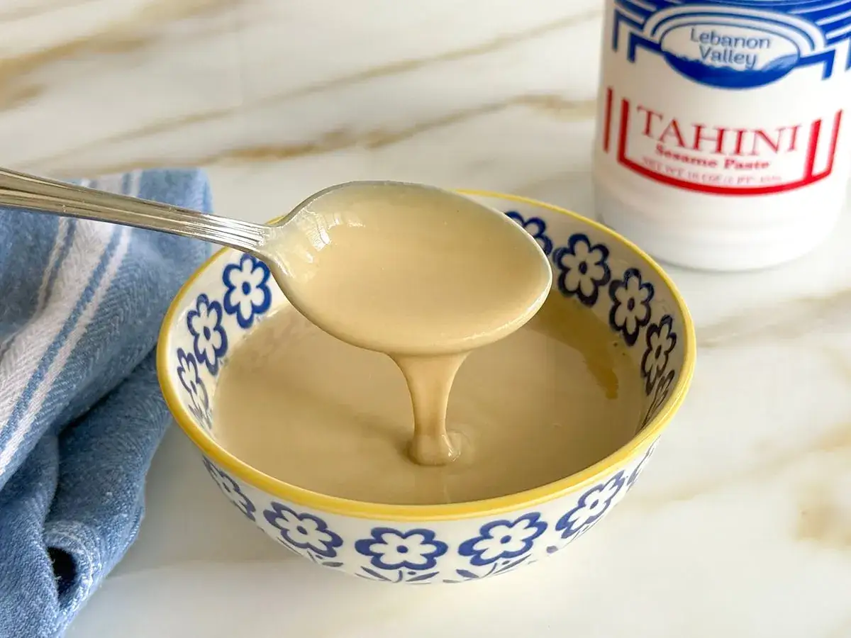 Ein L&ouml;ffel cremiges Tahin tropft in eine Sch&uuml;ssel. Was ist Tahin? Eine k&ouml;stliche Sesampaste, die in der K&uuml;che vielseitig einsetzbar ist.