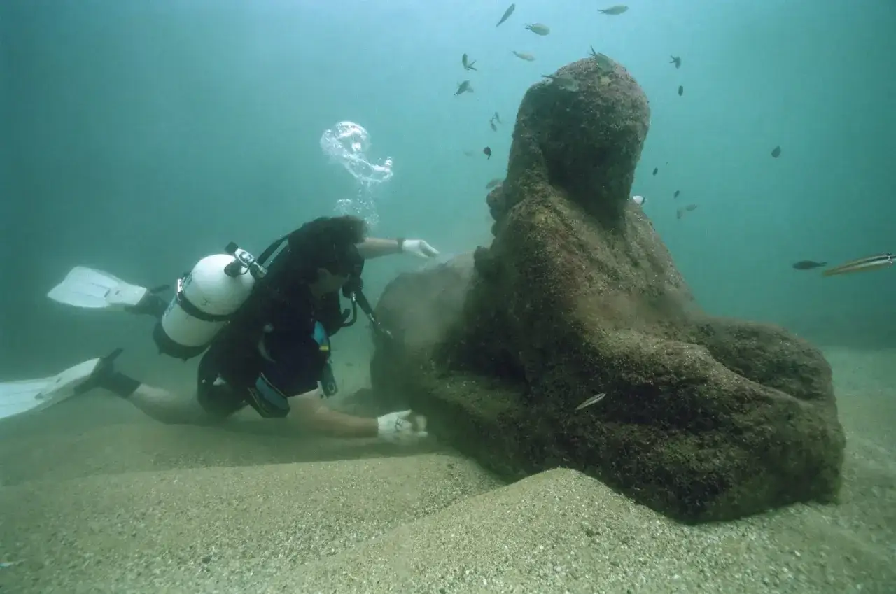Plongeur explorant une statue sous-marine, vestige du phare d'Alexandrie. Des poissons nagent autour de cette ruine antique.