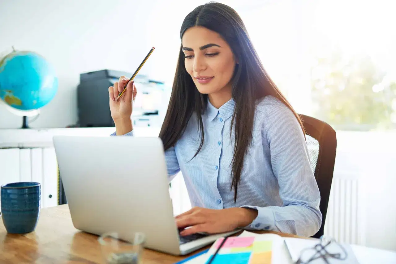 A woman thoughtfully considers her next steps in stress management coaching, holding a pencil while working on her laptop.