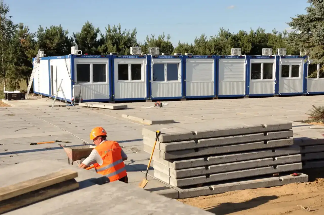 Budowlańcy przygotowują teren pod budowę, układając płyty drogowe betonowe. W tle kontenery biurowe.