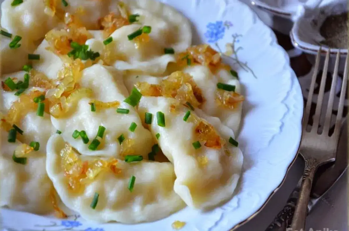 Pyszne pierogi z ziemniakami, okraszone skwarkami i szczypiorkiem, na białym talerzu z niebieskimi kwiatami.