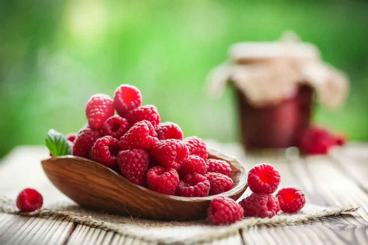 Frische Himbeeren in einer Holzschale, ein Symbol f&uuml;r gesunde Ern&auml;hrung. Im Hintergrund ein Glas Marmelade.