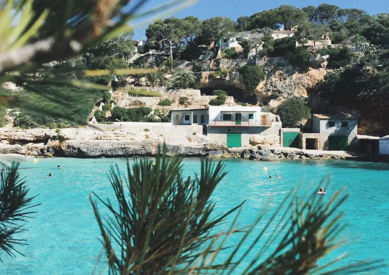 Traumhafter Urlaub auf Mallorca: Türkises Wasser, Felsenküste mit Häusern und Bäumen, Menschen schwimmen.