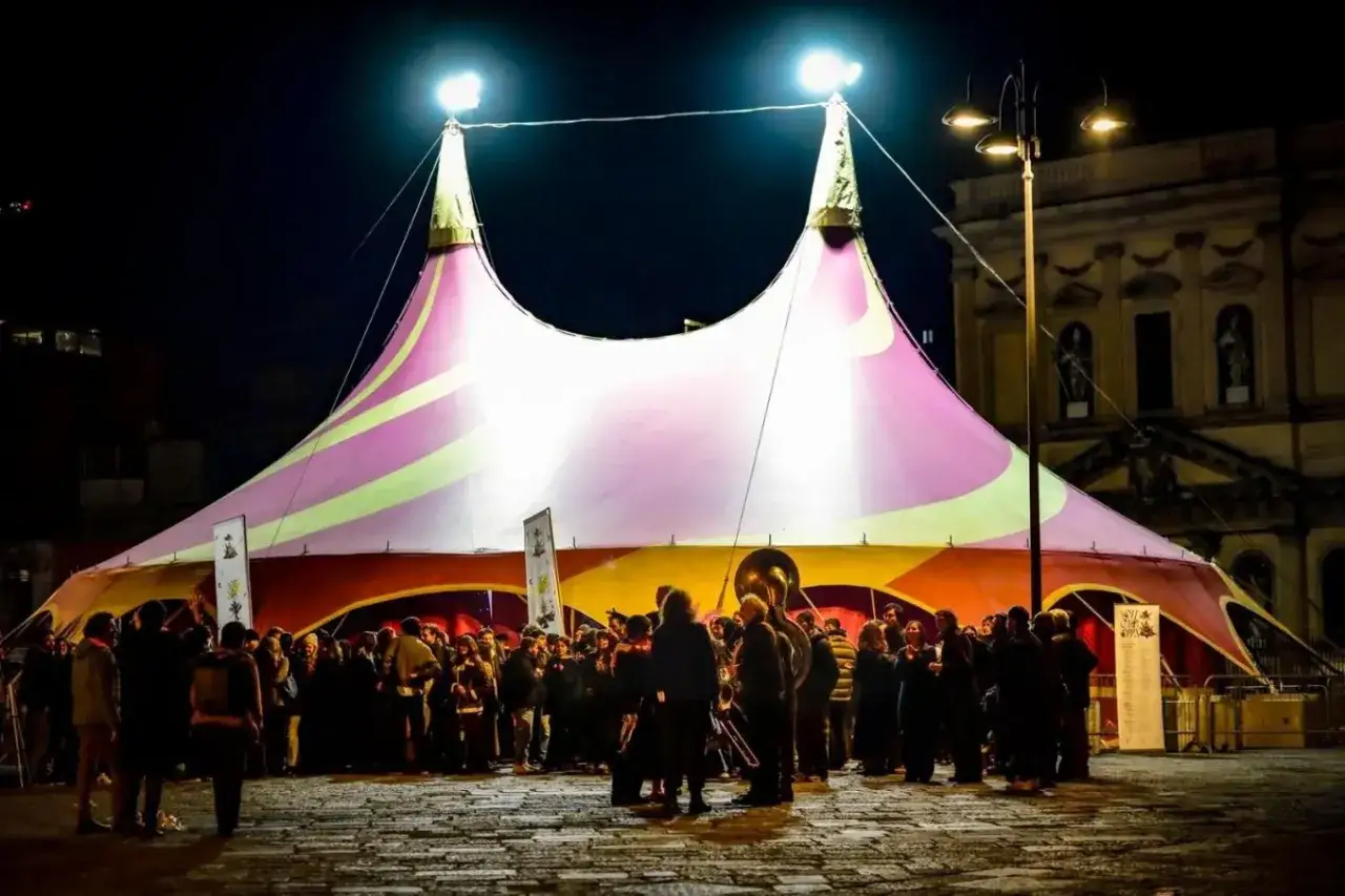 Tenda colorata illuminata di notte, con una folla radunata. Sembra un evento di carnevale, forse con un tocco di mistero, come se la morte stessa fosse invitata alla festa.