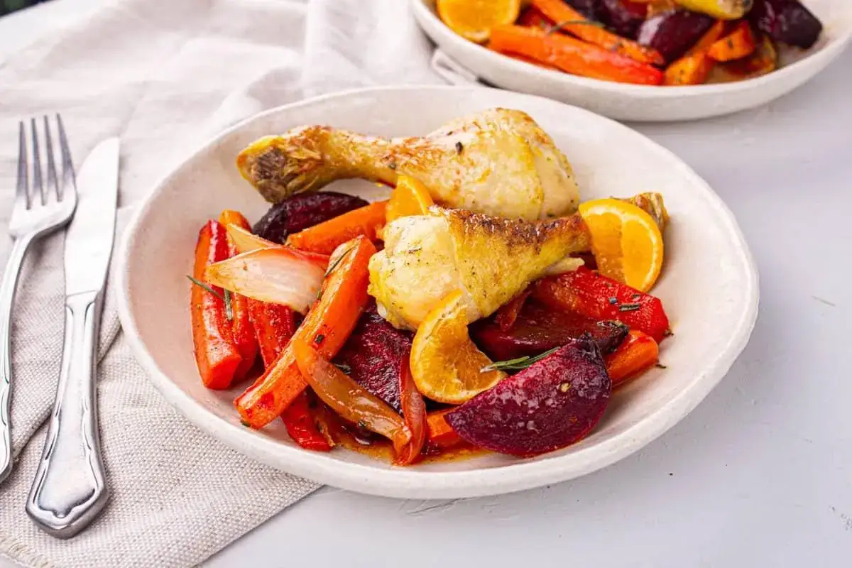 Un délicieux pilon de poulet rôti avec des carottes, des betteraves, des oignons et des quartiers d'orange, le tout servi dans une assiette blanche.