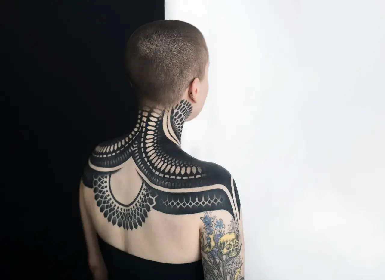 Una persona con un intrincado tattoo negro que cubre su cuello, hombros y espalda. El diseño es geométrico y orgánico, con detalles que recuerdan a escamas o plumas.