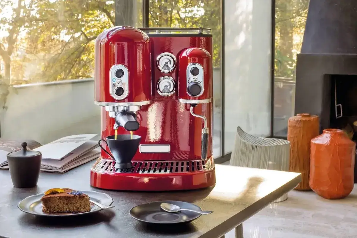 Macchina da caffè rossa lucida, con due bar, prepara un espresso. Sul tavolo, una fetta di torta e un libro.