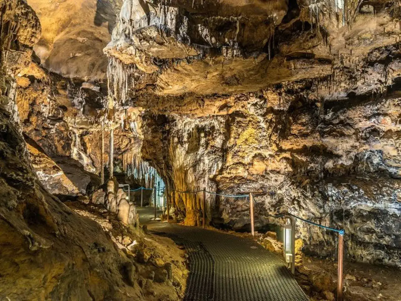 Interior de cuevas cerca de Madrid, con estalactitas y estalagmitas iluminadas, y un sendero met&aacute;lico para visitantes.
