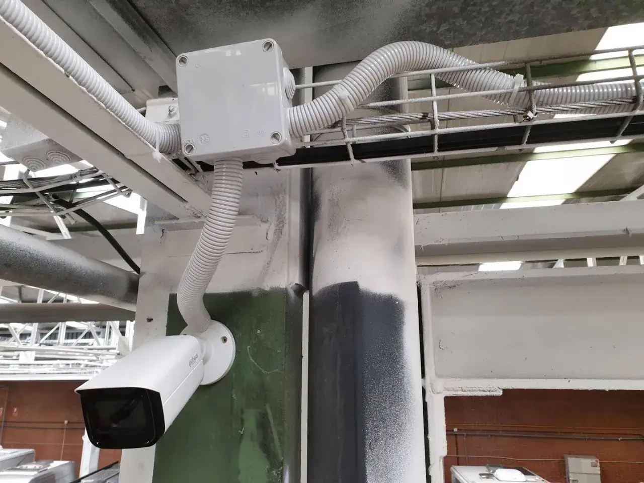 Cámara de circuito cerrado tv blanca, montada en una columna, con cables protegidos por tubos corrugados y una caja de conexiones.