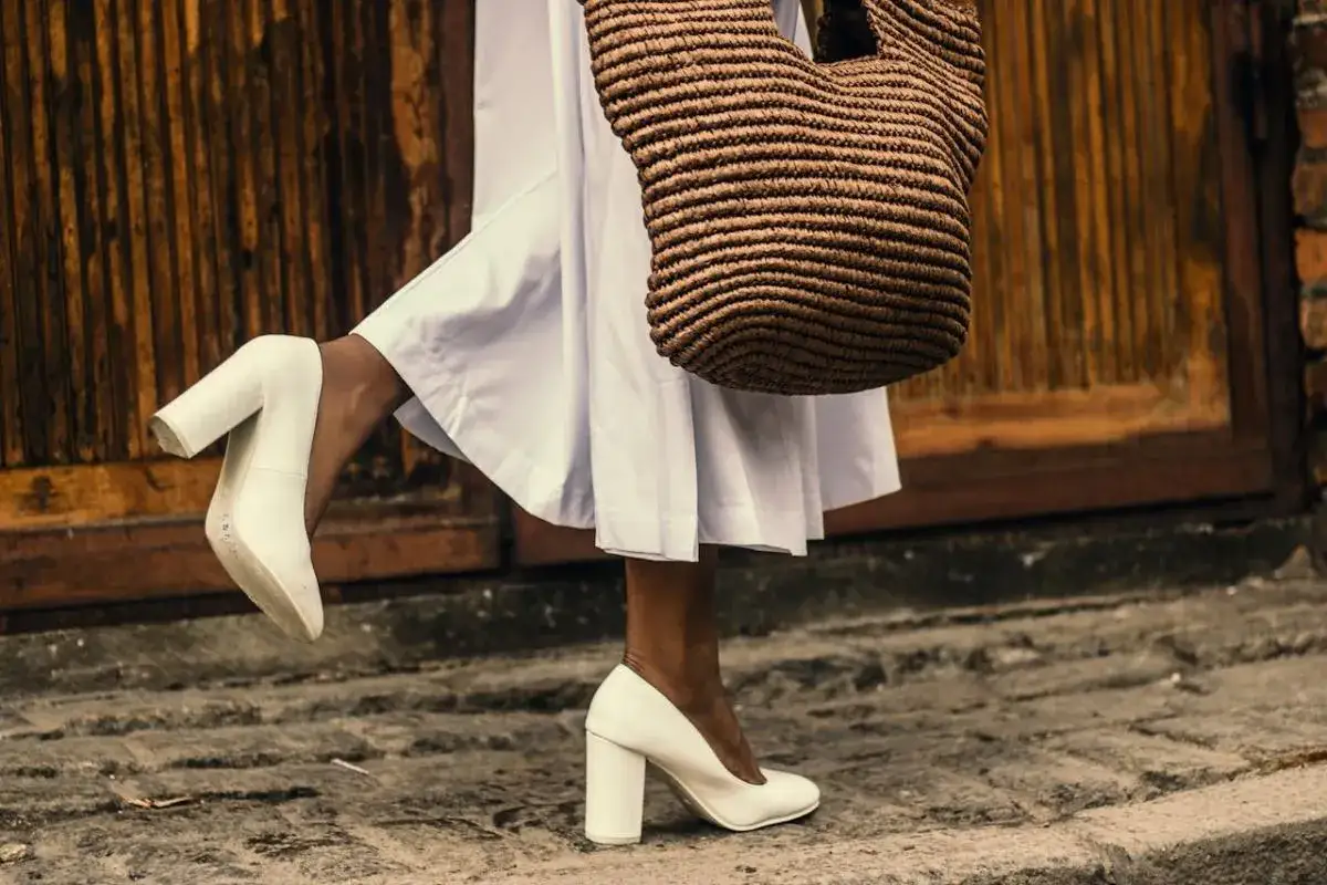 Białe, lakierowane czółenka na słupku to idealne buty do sukienki za kolano. Stylizacja uzupełniona plecioną torbą.