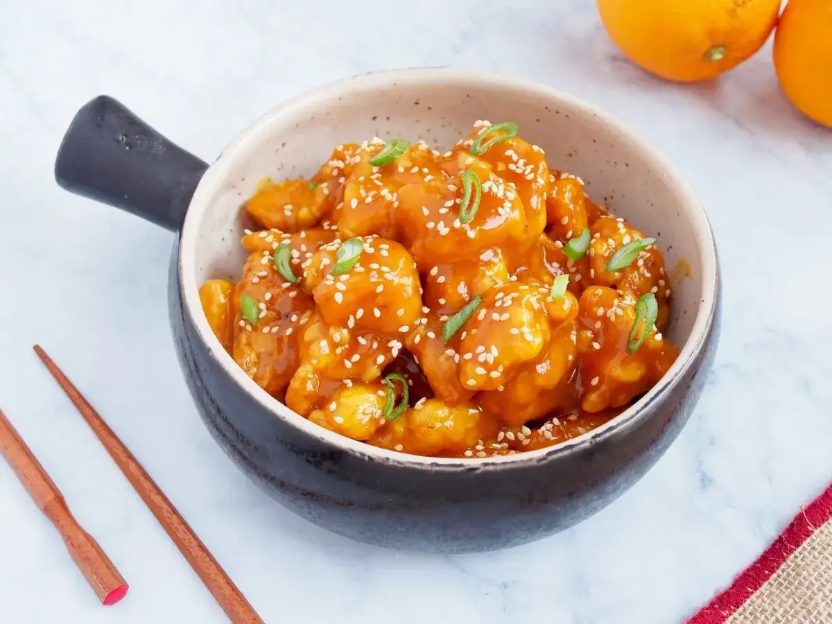 Un delicioso pollo a la naranja, cubierto de semillas de s&eacute;samo y cebolleta, servido en un cuenco con palillos de madera.