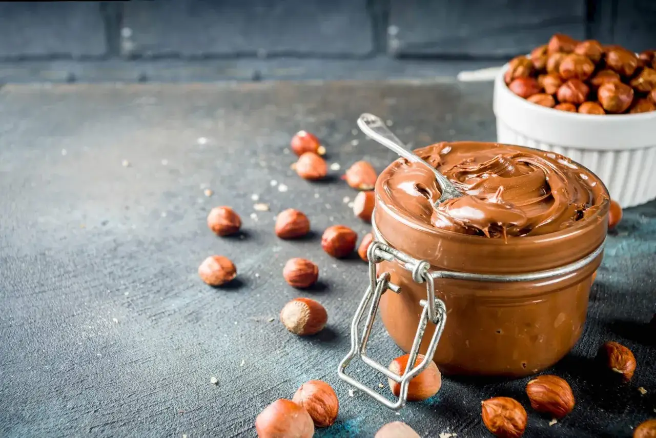 Crema di nocciole fatta in casa, cremosa e invitante, con nocciole sparse intorno. Un cucchiaino affonda nella deliziosa crema.