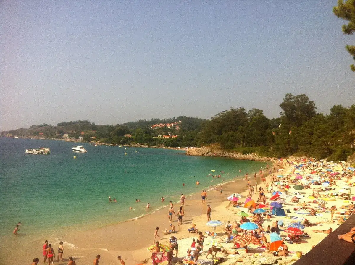 Gente disfrutando del sol y el mar en la concurrida playa Areacova. Barcos navegan en las aguas turquesas.