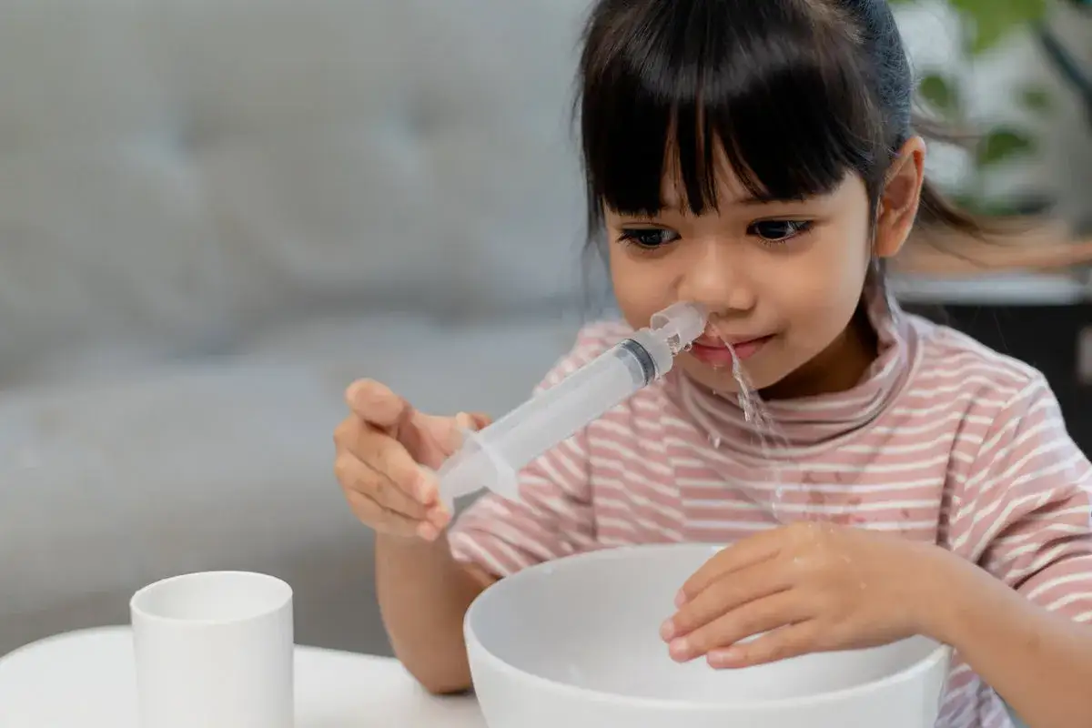 Niña realizando lavado nasal con jeringa, agua goteando en un cuenco.