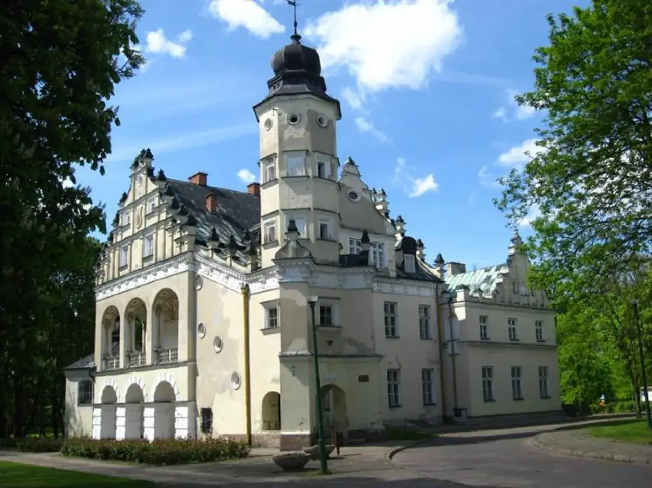 Poddębice pałac, zabytkowa budowla z wieżą i arkadowym gankiem, otoczona zielenią.