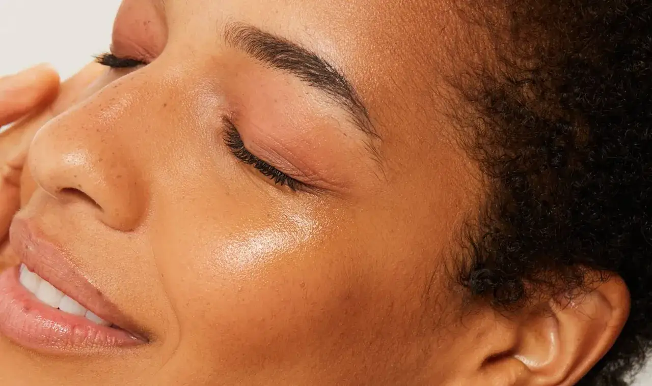 Close-up of a woman's face, showcasing glowing skin after enzyme exfoliation. Her eyes are closed, and a hand gently touches her cheek.
