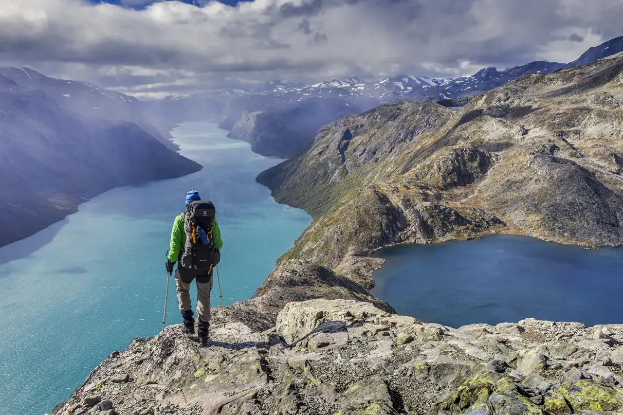 Vandrare med ryggsäck njuter av en hisnande utsikt över turkosa sjöar och snöklädda berg under en Jotunheimen vandring.
