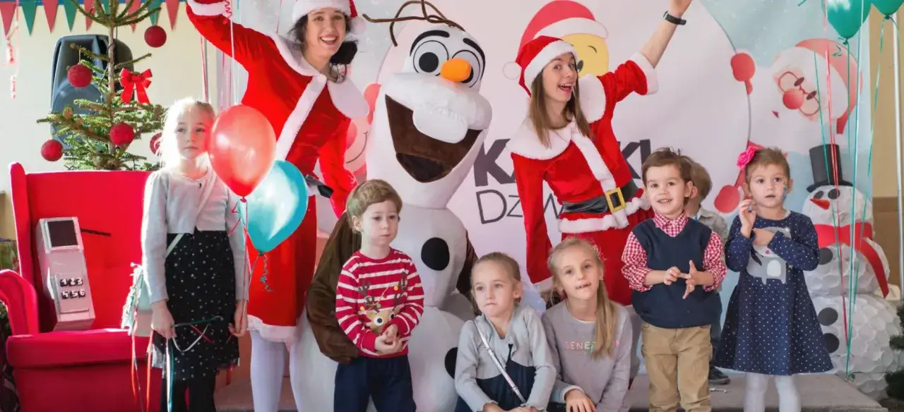 Radosne mikołajki w hotelu! Dzieci pozują z Paniami Mikołajkami i bałwanem Olafem, w tle choinka i balony.
