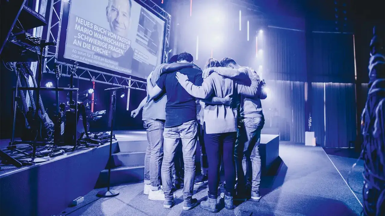Eine Gruppe von Menschen umarmt sich auf einer B&uuml;hne. Im Hintergrund wird ein neues Buch von Pastor Mario Wahnschaffe beworben, das Fragen an die Kirche stellt. Dies k&ouml;nnte eine Szene aus einem Gottesdienst von Pfingstlern sein.