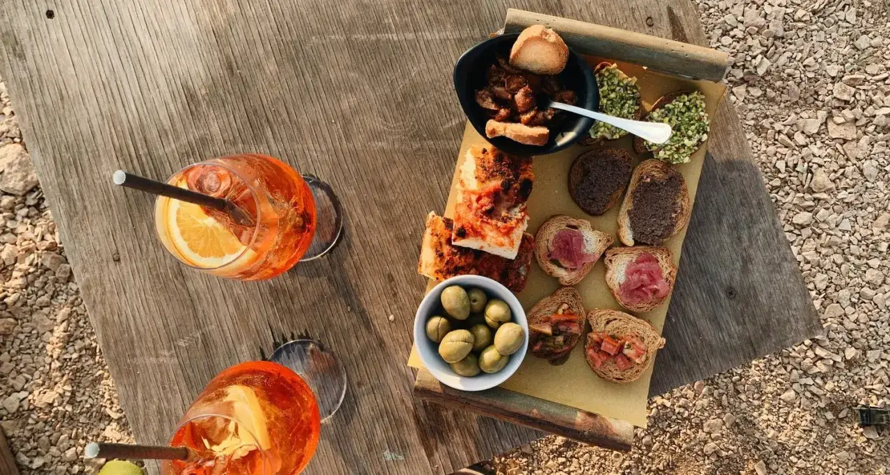 Två glas Aperol Spritz med apelsinskiva och sugrör, samt en bricka med bruschetta, oliver och andra tilltugg. Detta är vad aperitivo betyder – en härlig stund före maten.