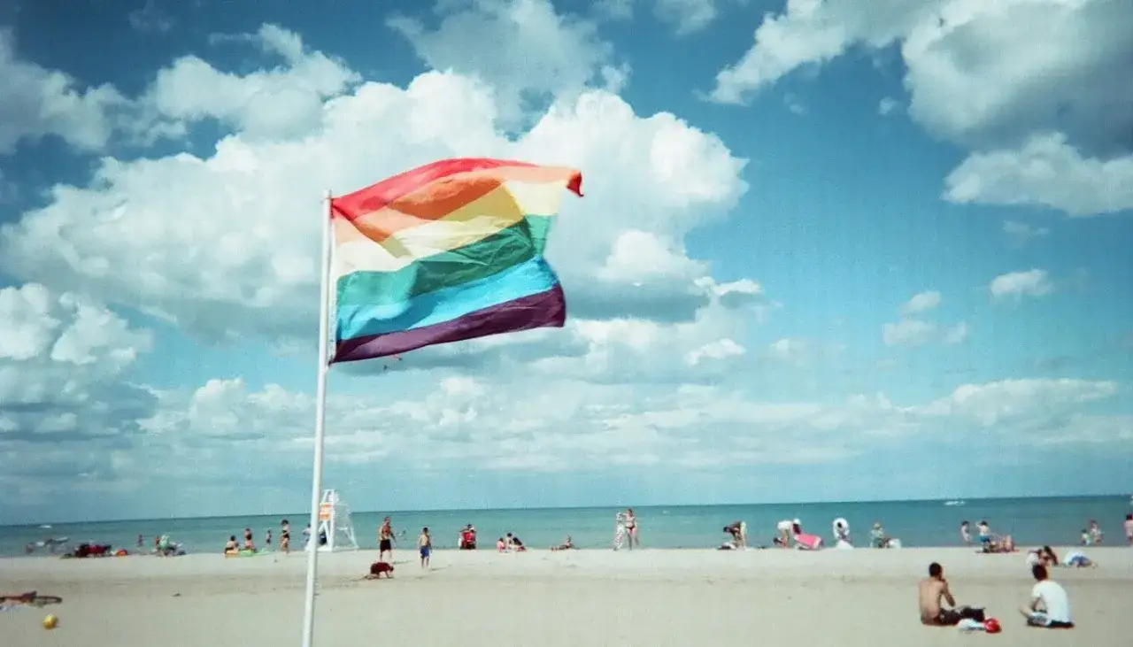 A rainbow flag flies over a sunny gay beach in Chicago. People relax on the sand by the blue lake under a cloudy sky.